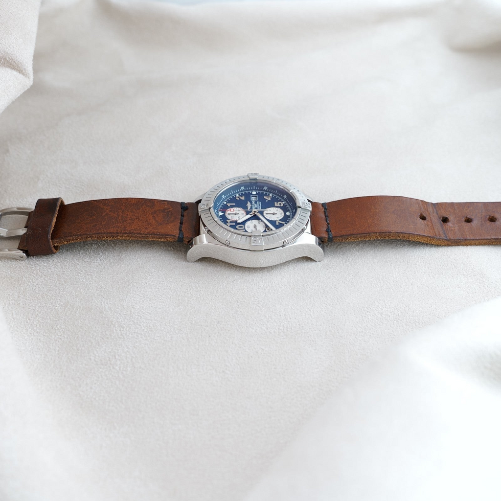 Wrist Flex Timepieces Breitling Super Avenger chronograph with blue dial and brown leather strap on a neutral fabric.